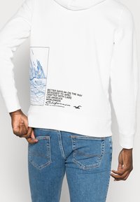 Sudadera blanca con un diseño gráfico azul en la parte posterior, que incluye un paisaje urbano y detalles de texto, con un corte relajado.