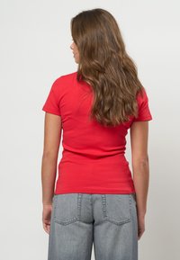 Pitkät, aaltoilevat ruskeat hiukset omaava nainen, jolla on päällään istuva punainen lyhythihainen paita ja vaaleanharmaat farkut, seisoo valkoista seinää vasten.