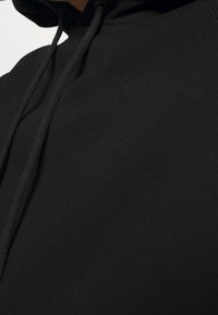 Svart huvtröja i mjukt material. Har en framficka och dragsnören, med ett slätt, texturerat tyg.