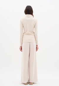 Bežinė megzta turtleneck tipo palaidinė su prigludusiu dizainu, derinama su šviesiai bežinėmis plačiomis kelnėmis. Abu drabužiai pasižymi švelnia tekstūra.