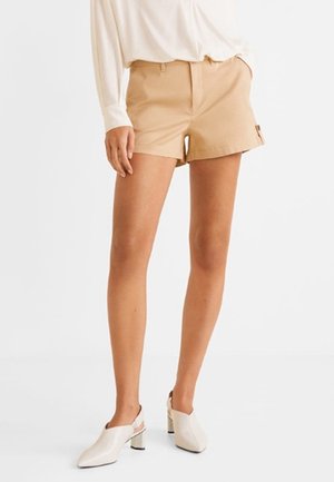 Kvinde iført beige skræddersyede shorts, hvid langærmet top og hvide slingback sko med hæl, stående med den ene hånd i lommen.