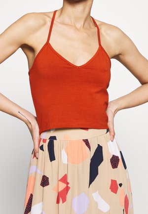 Femme portant un haut court rouge sans manches avec des bretelles fines et une jupe beige à motifs abstraits multicolores, mains posées sur les hanches.