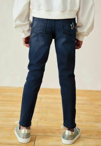 Sötétkék, slim fit fazonú farmernadrág, öt zsebes kivitelben, a hátsó zseben virágmintás hímzéssel. Csillogó cipővel párosítva.