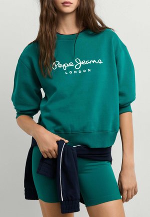 Giovane donna che indossa una felpa verde Pepe Jeans London e pantaloncini coordinati, tiene una giacca blu navy legata intorno alla vita.