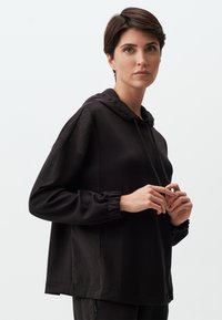 Sweatshirt à capuche noire en tissu doux avec des poignets élastiques, un cordon de serrage et une coupe décontractée, comportant des coutures minimales et sans motifs.