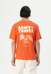 Korte mouw oranje t-shirt met witte bedrukte tekst en afbeeldingen van palmbomen, een zon en een strandparasol op de achterkant.