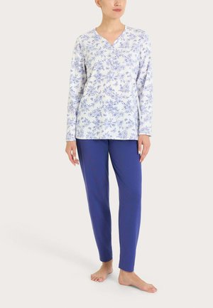 Donna che indossa una maglia bianca a maniche lunghe con stampa floreale blu e pantaloni blu alla caviglia, in piedi a piedi nudi su uno sfondo neutro.