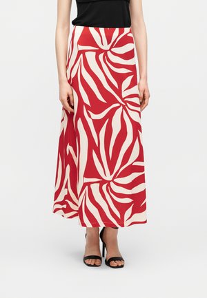 Donna con una gonna lunga a motivo astratto rosso e bianco, top nero senza maniche e sandali neri con tacco alto, in piedi davanti a uno sfondo bianco.