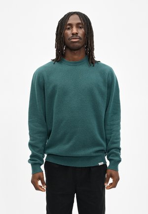 Man met lange dreadlocks, gekleed in een teal lange-mouwen trui en zwarte broek, staand tegen een effen witte achtergrond.