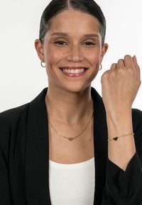 Gold jewelry set features a twisted chain necklace and a bracelet with heart-shaped accents. Model wears a black blazer and white top.