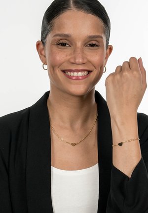 Das Goldschmuckset besteht aus einer gedrehten Ketten-Halskette und einem Armband mit herzförmigen Akzenten. Das Model trägt einen schwarzen Blazer und ein weißes Oberteil.
