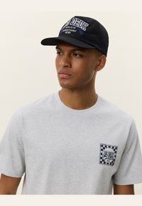 Grå T-skjorte med kort erm og brystlomme som har et rutete mønster og "LES DEUX" grafikk. Svart baseballcaps med brodert tekst.