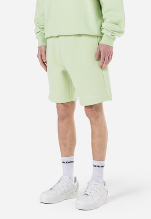 Hellgrüne Baumwollshorts mit lockerem Schnitt, Seitentaschen und kleinem Logo am Saum. Kombiniert mit weißen Sneakers und gestreiften Socken.