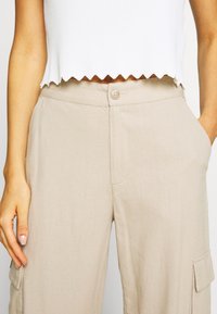 Beige cargo pants made of lightweight fabric, featuring a button closure, side pockets, and a relaxed fit. Scalloped white crop top above.