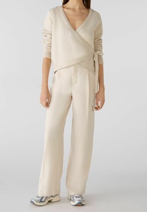 Kvinde iført beige omslagsstrik med sidesløjfe, vidde cremefarvede bukser og hvide-grå sneakers stående mod en ensfarvet baggrund.