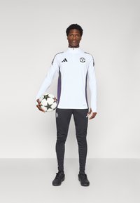 Hvit jakke med glidelås for trening, med lilla og svarte detaljer, kombinert med svarte treningbukser, holder en fotball, iført svarte sko.