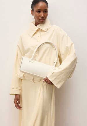 Žena u bež širokoj jakni i suknji drži malu bijelu pravokutnu torbicu s zaobljenom drškom.