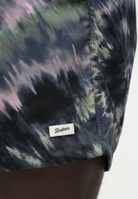 Κοντινή λήψη μαγιό σορτς batik με πράσινους, μαύρους και μοβ τόνους, που διαθέτουν μικρή λευκή ετικέτα με την ένδειξη "Bather" κοντά στο τελείωμα.
