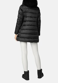 Schwarzer Steppmantel mit einem Kunstpelzkragen, mit einem gesteppte Design, knielang geschnitten, kombiniert mit weißen Leggings und schwarzen Stiefeletten.