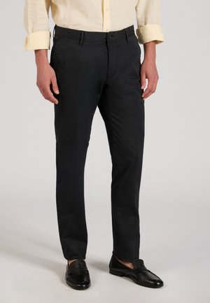 Mężczyzna ubrany w czarne spodnie o prostych nogawkach, czarne buty wsuwane oraz beżową koszulę z długim rękawem i podwiniętymi mankietami.