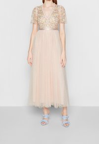 Lungo vestito in tulle color blush con ricami floreali, scollatura a V e maniche a sbuffo. Abbinato a sandali intrecciati blu chiaro.