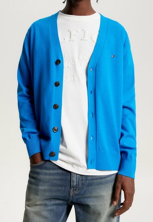 Cardigan bleu en tissu doux, avec un col en V, sept grands boutons et des poignets côtelés. Porté sur un t-shirt blanc.