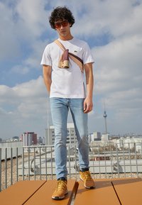 Tricou alb, blugi de culoare bleu deschis și pantofi bej. O geantă maro și bej purtată pe piept. Ochelari de soare portocalii intens. Cadru urban pe acoperiș.