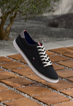 Schwarze Leinenschuhe mit weißen Schnürsenkeln und Sohle, die ein kleines rot-weiß-blaues Logo tragen, auf Holz- und Kieselbodenfliesen präsentiert.