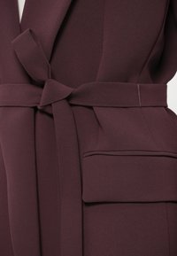 Bordó átlapolós ruha kötött övvel, sima textúrával és elülső zsebbel, tiszta vonalakkal és szabott tervezési elemekkel.