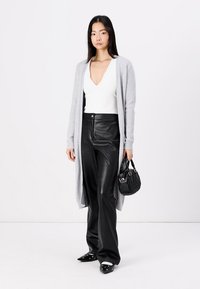Langer grauer Cardigan über einem weißen V-Ausschnitt-Top, schwarze Leder-Hosen mit hoher Taille und schwarze Handtasche mit gestepptem Muster. Spitz zulaufende Slipper.