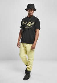 Sort bomulds T-shirt med guldskrift, gule sweatpants og sort spandhat. Sorte sneakers fuldender outfittet, der har en skuldertaske.