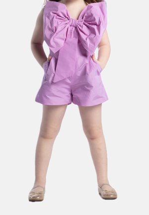 Combi-short lilas avec un grand nœud au niveau du col, comportant deux poches latérales et une texture lisse et brillante. Porté avec des chaussures plates dorées.