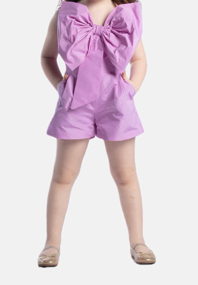 Fioletowy kombinezon z dużą kokardą przy dekolcie, z dwiema bocznymi kieszeniami i gładką, błyszczącą fakturą. Noszony z złotymi płaskimi butami.