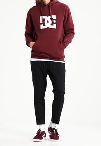 Felpa bordeaux con logo DC bianco, tasca a canguro, abbinata a pantaloni neri a sigaretta e sneakers bordeaux con dettagli bianchi.