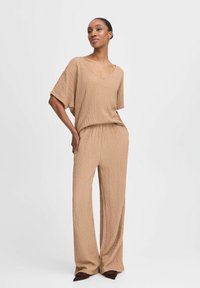 Beige, strukturiertes Zweiteiler-Outfit mit einem locker sitzenden Oberteil und weitem Hosenbein. V-Ausschnitt und elastischer Taillenbund für zusätzlichen Komfort. Braune Schuhe.