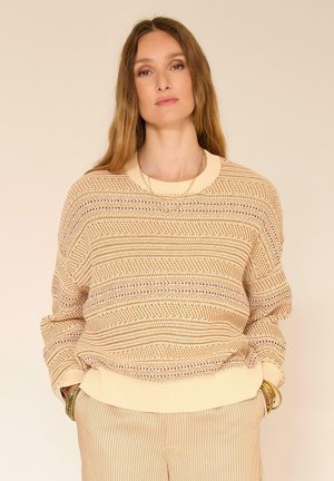 Femme portant un pull en maille beige à motifs et un pantalon plissé clair, les mains dans les poches et des bijoux discrets, sur un fond neutre.