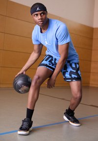 Modré sportovní tričko s logem, černé a modré vzorované šortky, černé tenisky s oranžovými akcenty, držící černý basketbal v tělocvičně.