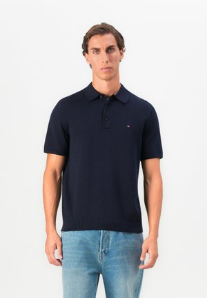 Nuori mies, jolla on yllään tummansiniset lyhythihainen poolopaita ja vaaleansiniset farkut, seisoo plain-valkoisen taustan edessä.