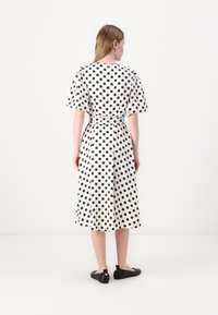 Vestido branco com bolinhas pretas, midi, com mangas curtas bufantes, cintura ajustada e um corte solto. Usado com sapatilhas pretas.