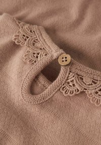 Top de punto en rosa suave con ribete de encaje y detalle de botón de madera. La tela tiene una textura fina y un diseño sutilmente estampado.