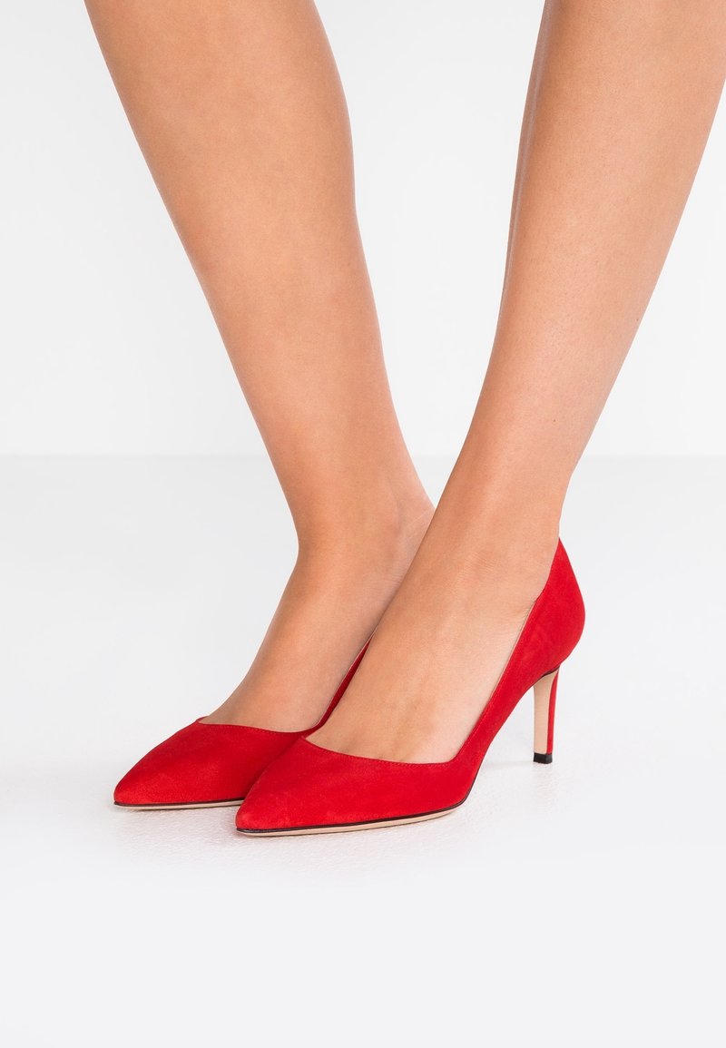 Jambes portant des escarpins en daim rouge vif à bout pointu avec des talons aiguilles fins sur un sol blanc uni.