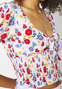 Blusa in tessuto leggero con stampa floreale caratterizzata da fiori rossi, viola e gialli su sfondo bianco, con arricciature in vita e maniche a palloncino.