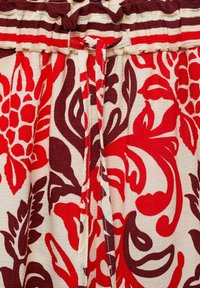 Bloemenpatroon stof in rood en donker bordeaux op een crème achtergrond. Beschikt over een gerimpelde tailleband en gestrikte accenten. Gelaagd weefsel.