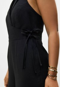 Combinaison noire avec un corsage ajusté, un design enveloppant et un détail de nœud sur le côté, comportant des coutures verticales et une texture lisse. Bracelets dorés au poignet.