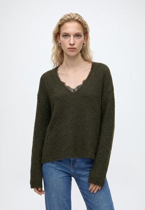 Jeune femme aux cheveux blonds ondulés porte un pull vert foncé col en V par-dessus un top en dentelle et un jean bleu, debout devant un fond uni.