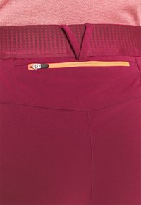 Leggings de sport marron rouge avec une large ceinture ornée d'un motif texturé et une petite poche zippée orange à l'arrière. Finition du tissu lisse.