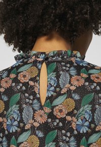 Sort blomsterbluse med flæsekant ved halsen og en nøglehulssnit bagpå. Stoffet er prydet med blomster i blå, orange og grøn.