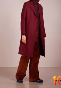 Abrigo de mezcla de lana burdeos con cuello de muesca y botones de doble botonadura, combinado con pantalones de pana marrón de pernera ancha y botas negras.