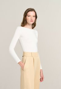 White long-sleeve top with a boat neckline, paired with high-waisted beige trousers featuring side pockets and pleated design. Smooth fabric texture.