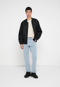 Juoda bombonešio striukė, šviesiai smėlio spalvos marškinėliai su logotipu, šviesiai mėlynos džinsinės kelnės ir juodi sportbačiai. Paprastas dizainas su laisvu siluetu ir lygiu audiniu.